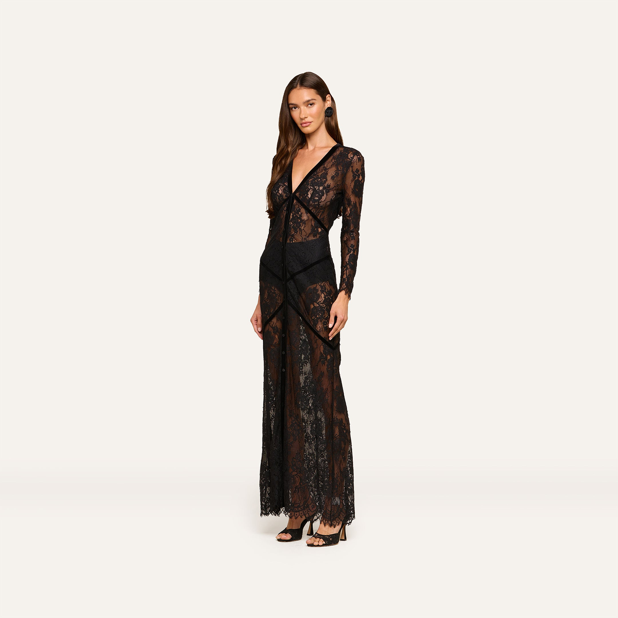 A woman poses in the Poesy Lace Maxi Dress, a black maxi-length gown featuring long sleeves and paired with black heels, set against a plain light background.