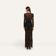 A woman stands facing away, wearing the Poesy Lace Maxi Dress—a black lace maxi-length dress with sheer panels and long sleeves—revealing a black bodysuit underneath. She has straight brown hair and is wearing black heels against a plain, light background.