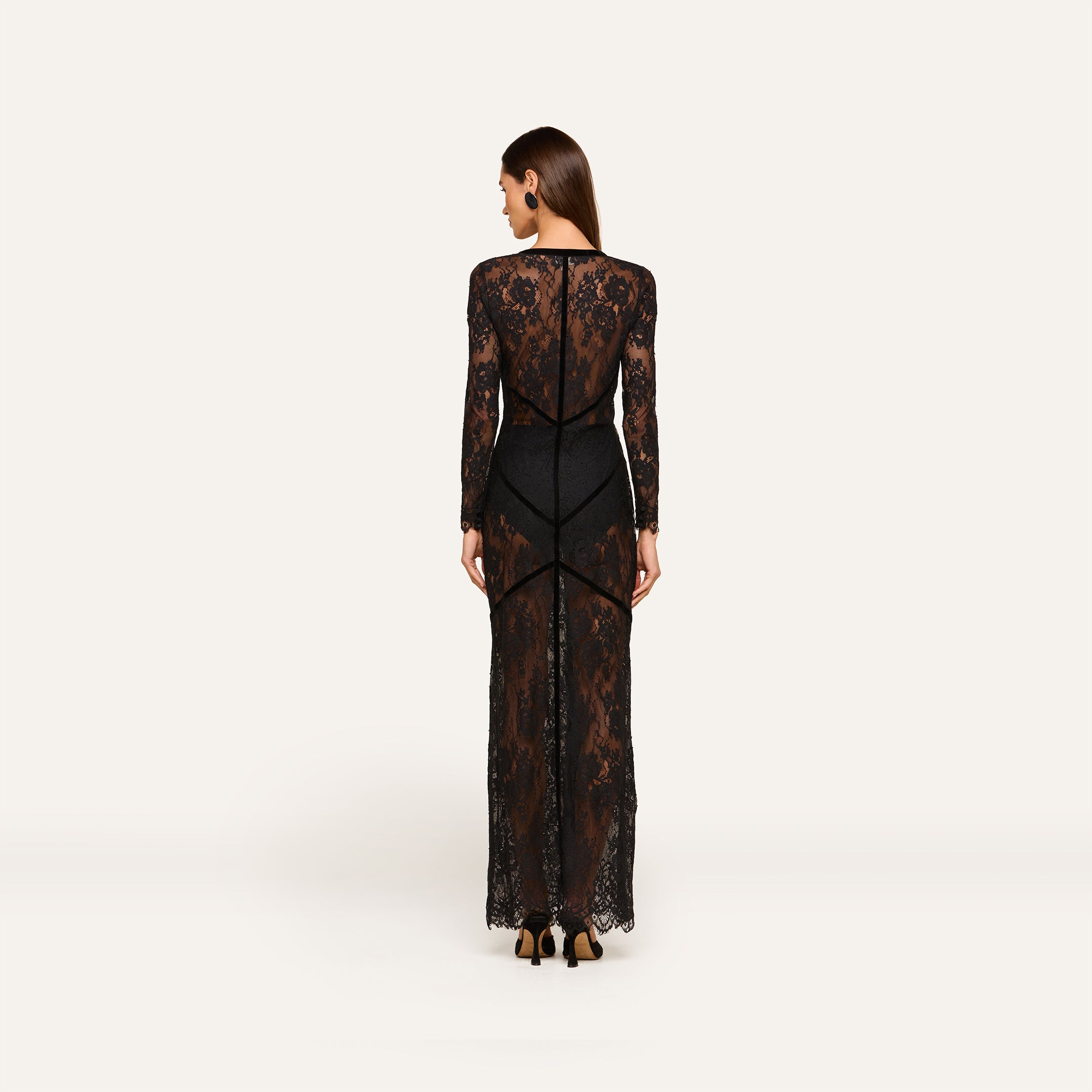 A woman stands facing away, wearing the Poesy Lace Maxi Dress—a black lace maxi-length dress with sheer panels and long sleeves—revealing a black bodysuit underneath. She has straight brown hair and is wearing black heels against a plain, light background.