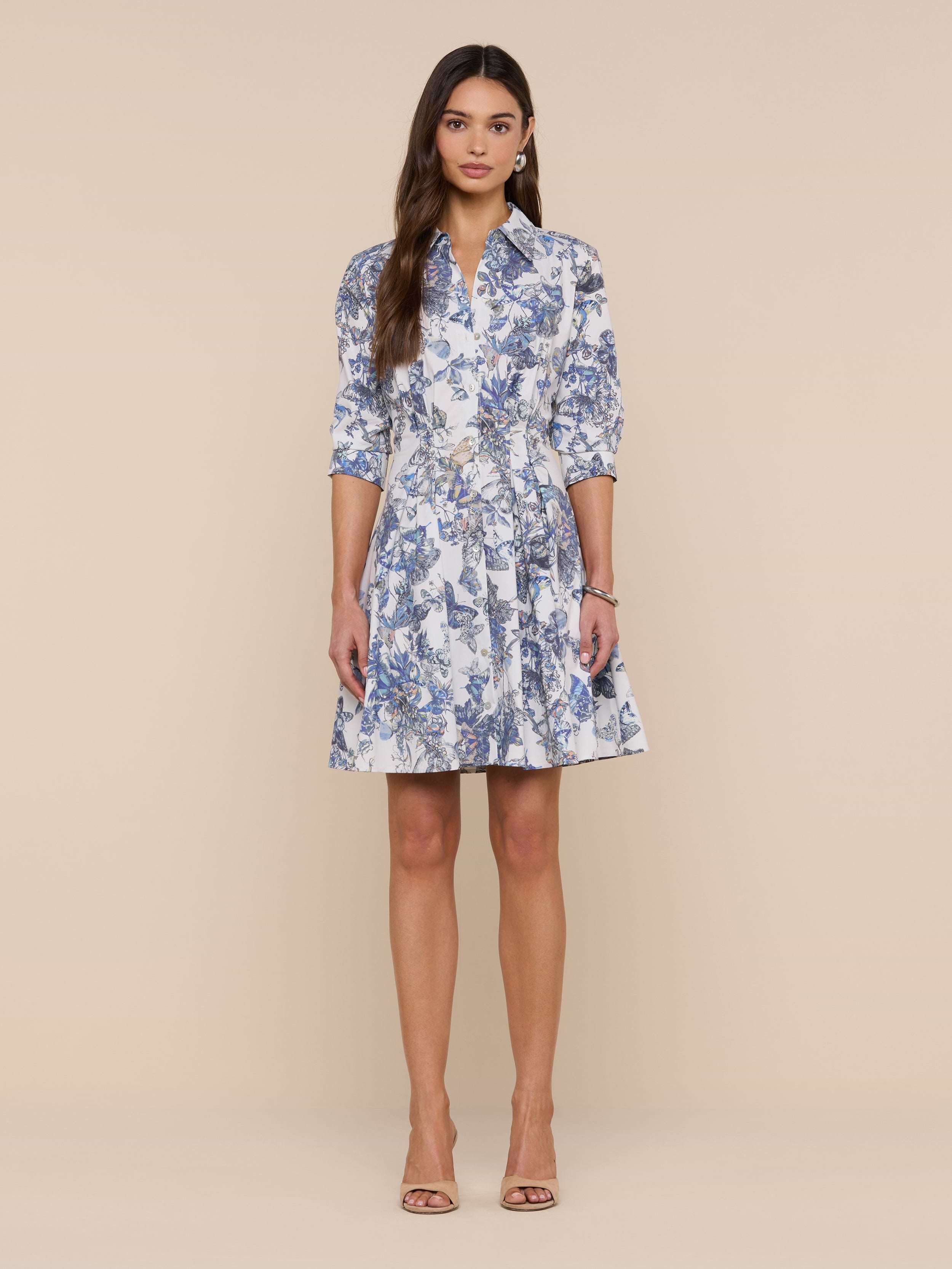 A woman stands against a plain background, wearing the Journi Shirt Dress—a knee-length, blue and white floral stretch cotton-blend shirt dress with three-quarter sleeves—paired with beige open-toe heels. Her long, straight hair is down, and her arms are relaxed by her sides.