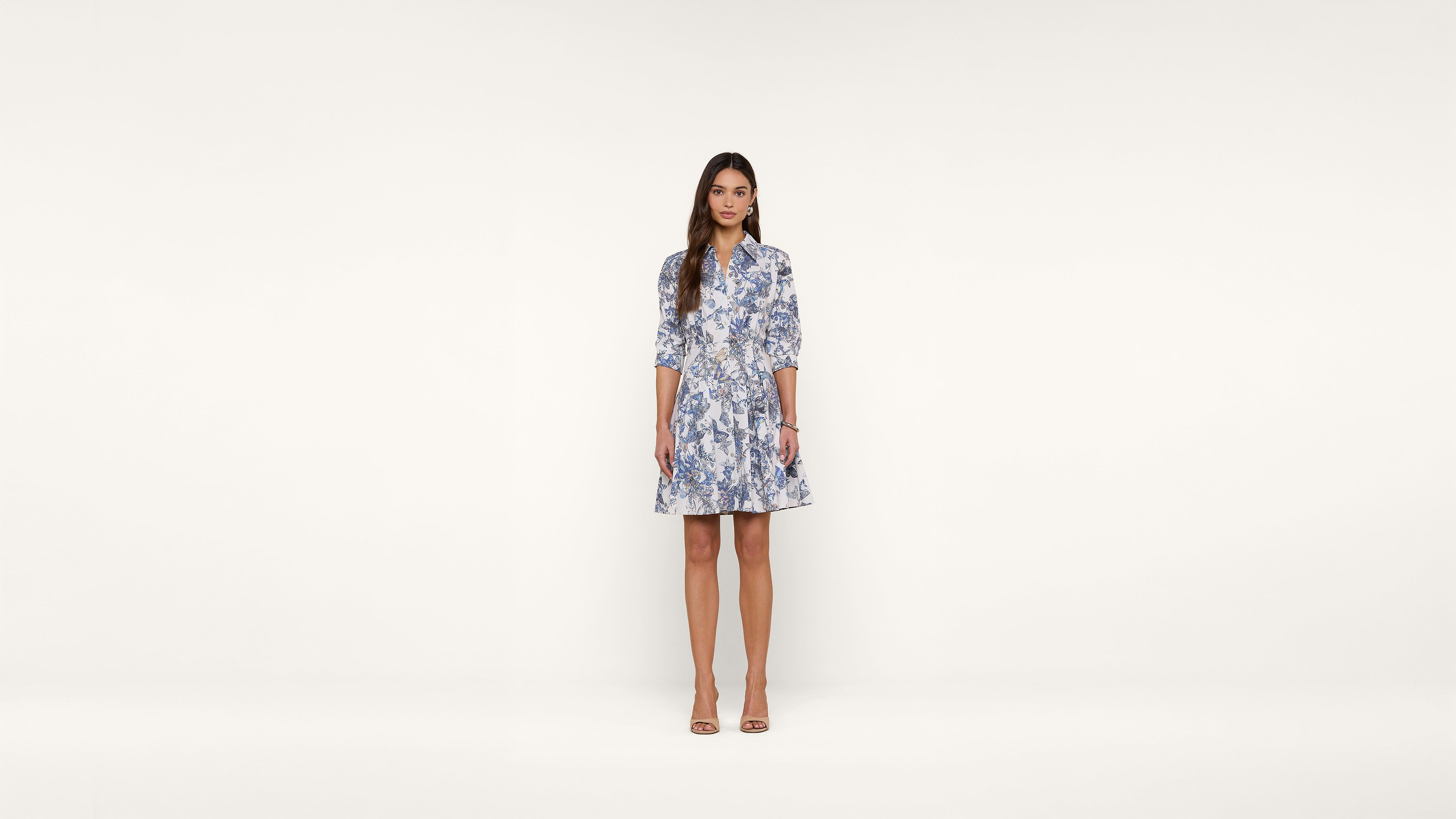 A woman stands against a plain white background wearing a light blue and white floral dress with three-quarter sleeves and beige heels.