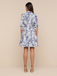 A woman stands facing away, wearing the Journi Shirt Dress—a knee-length design featuring blue and white floral and butterfly print patterns with three-quarter sleeves—paired with beige heels, set against a plain white background.