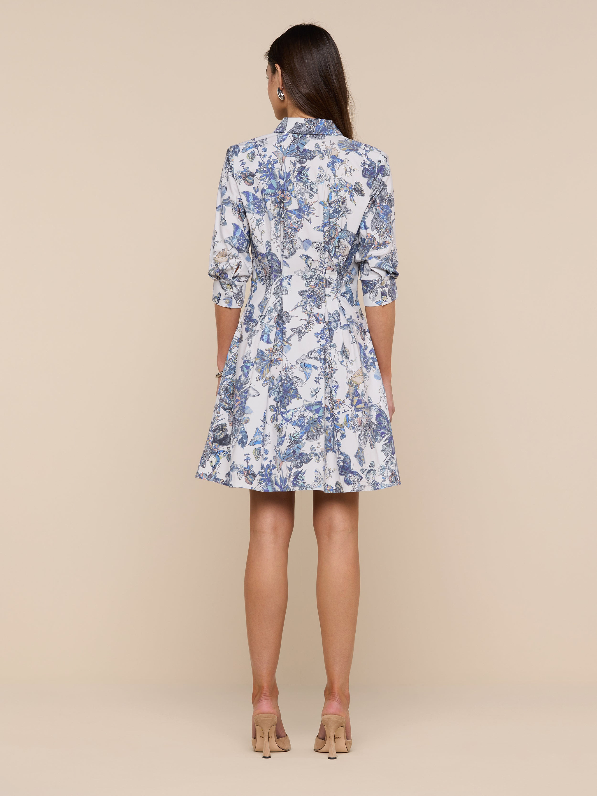 A woman stands facing away, wearing the Journi Shirt Dress—a knee-length design featuring blue and white floral and butterfly print patterns with three-quarter sleeves—paired with beige heels, set against a plain white background.