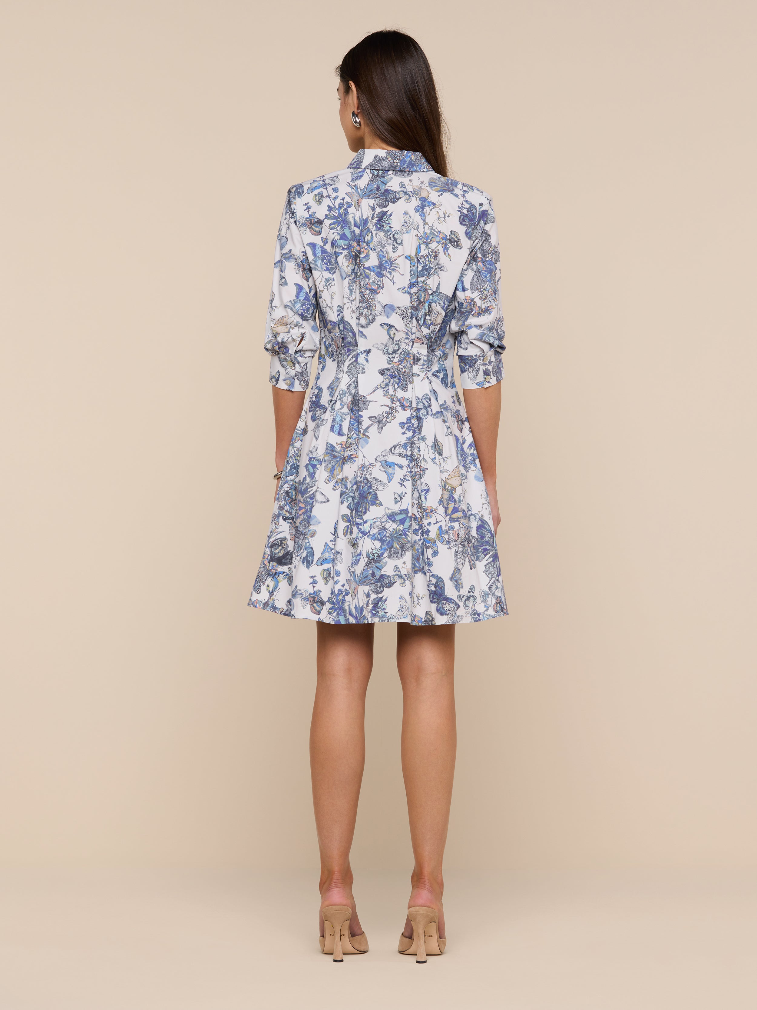 A woman stands facing away, wearing the Journi Shirt Dress—a knee-length design featuring blue and white floral and butterfly print patterns with three-quarter sleeves—paired with beige heels, set against a plain white background.