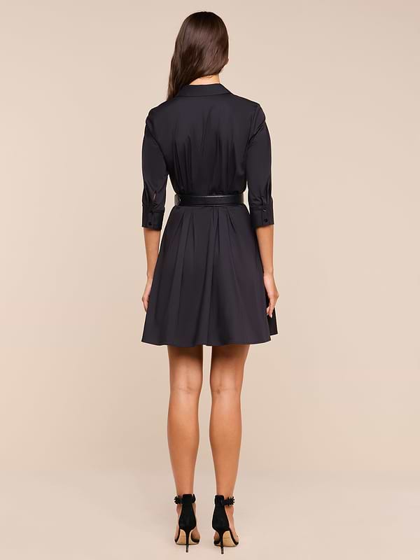 A woman with long brown hair stands facing away, wearing the Journi Shirt Dress—a black cotton-blend poplin shirt dress with a belt—and black high-heeled shoes against a plain white background.