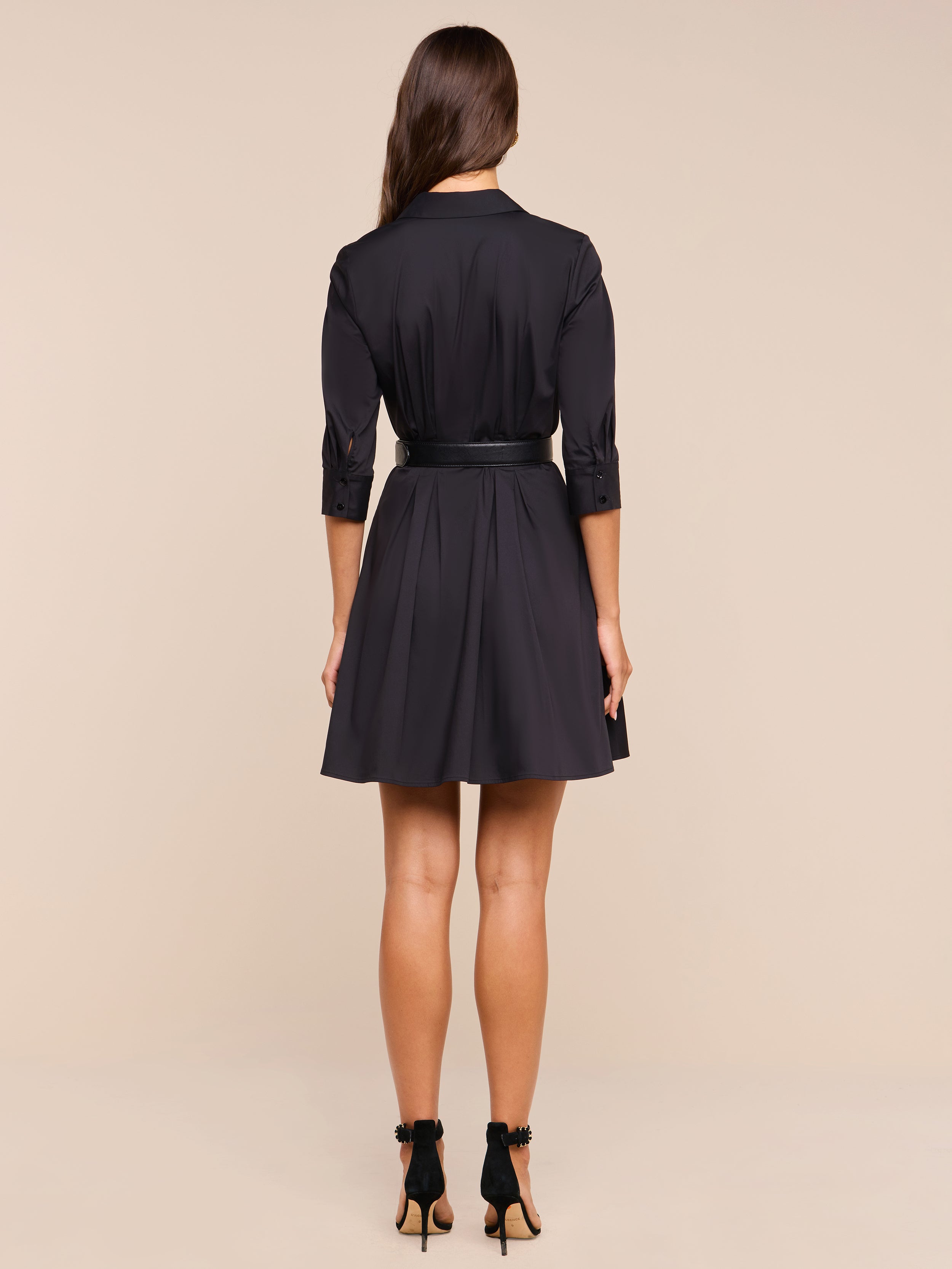 A woman with long brown hair stands facing away, wearing the Journi Shirt Dress—a black cotton-blend poplin shirt dress with a belt—and black high-heeled shoes against a plain white background.