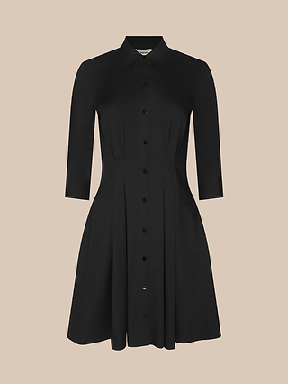The Journi Shirt Dress, designed in black cotton-blend poplin with a knee-length cut, showcases three-quarter sleeves, a collared neckline, and a pintuck waist for a flattering silhouette, displayed against a neutral beige background.