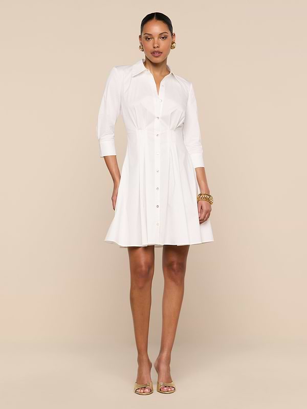A woman stands against a plain white background wearing the Journi Shirt Dress—a white, button-down, knee-length design with three-quarter sleeves. She accessorizes with gold jewelry and open-toe heels, her left hand in her pocket to highlight the timeless silhouette of this cotton-blend poplin piece.