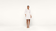 A woman stands against a plain white background wearing a white, button-down dress with three-quarter sleeves and beige heels. She has her hair pulled back and is accessorized with earrings and bracelets.
