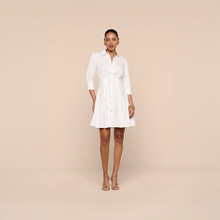 A woman stands facing forward against a beige background, wearing the Journi Shirt Dress—a classic white shirt dress with three-quarter sleeves—styled with gold hoop earrings, bracelets, and open-toe heels.