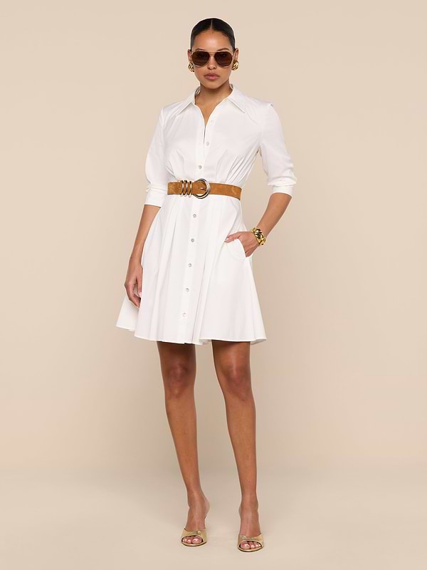 A woman wearing the Journi Shirt Dress—crafted from a white cotton-blend poplin—paired with a brown belt, sunglasses, and open-toed heels stands against a plain white background, one hand tucked in her pocket.