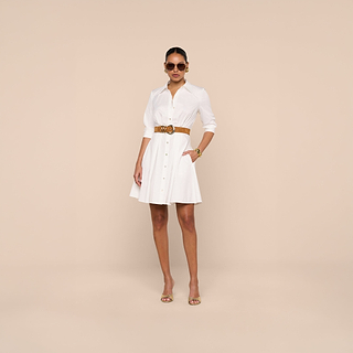 A woman stands against a beige background wearing sunglasses and the Journi Shirt Dress—a white stretch cotton-blend design finished with a brown belt—paired with gold heels and a gold bracelet. With one hand in her pocket, she exudes confidence.