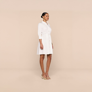 A woman stands in a neutral studio, wearing the Journi Shirt Dress—a white, three-quarter sleeve shirt dress crafted from a stretch cotton-blend fabric that complements its classic silhouette. She pairs it with beige heels, her hair styled neatly in a bun against a light beige background.