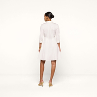 A woman stands on a white background, facing away. She wears the Journi Shirt Dress—a white, knee-length shirt dress with three-quarter sleeves crafted from cotton-blend poplin for a timeless silhouette. Her light-colored heeled shoes complement the look, and her hair is styled in a neat bun.