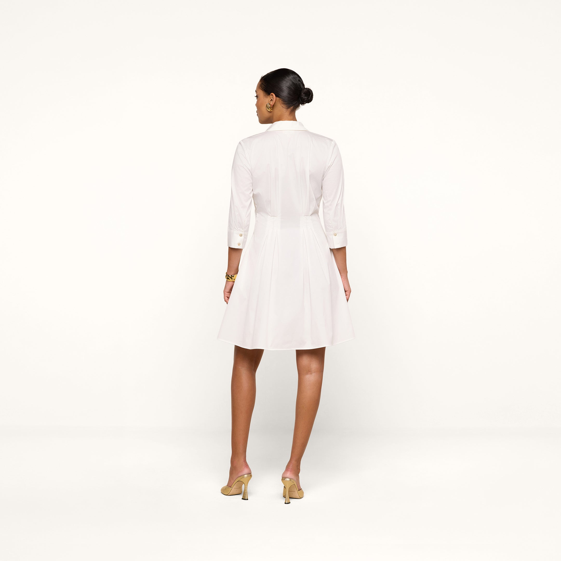 A woman stands on a white background, facing away. She wears the Journi Shirt Dress—a white, knee-length shirt dress with three-quarter sleeves crafted from cotton-blend poplin for a timeless silhouette. Her light-colored heeled shoes complement the look, and her hair is styled in a neat bun.