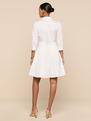 A woman, with her hair in a neat bun, faces away wearing the Journi Shirt Dress—white, knee-length with three-quarter sleeves—and beige heeled sandals, set against a beige background.