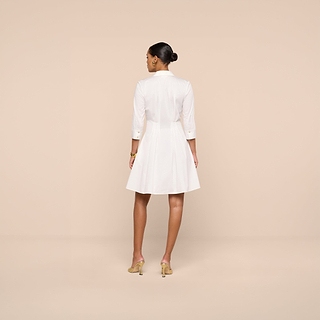 A woman stands facing away, wearing the Journi Shirt Dress—a white stretch cotton-blend design with three-quarter sleeves—paired with beige heels, set against a plain beige background.