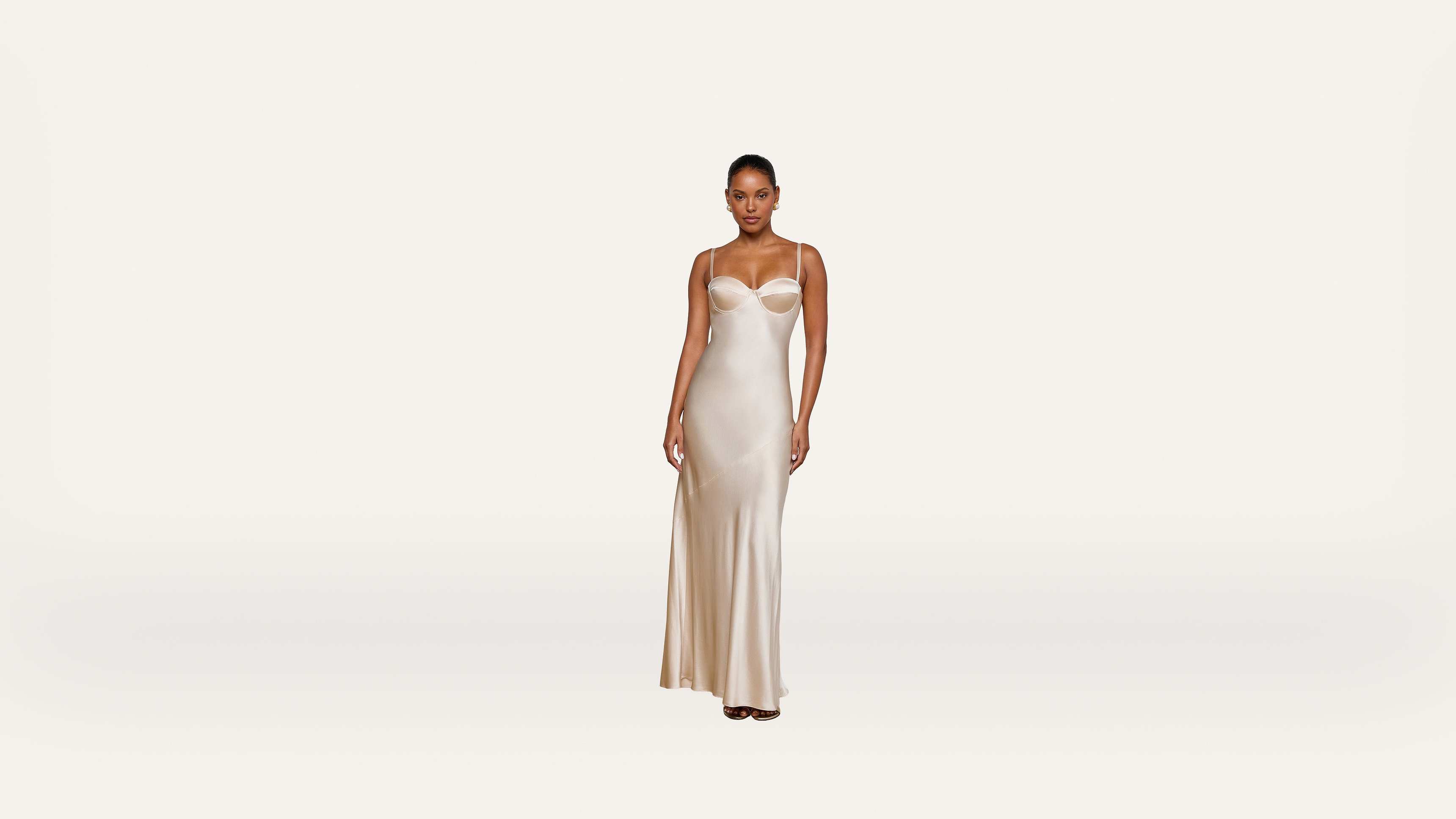 A woman wearing a long, cream-colored satin slip dress stands against a plain white background, facing forward with a neutral expression.