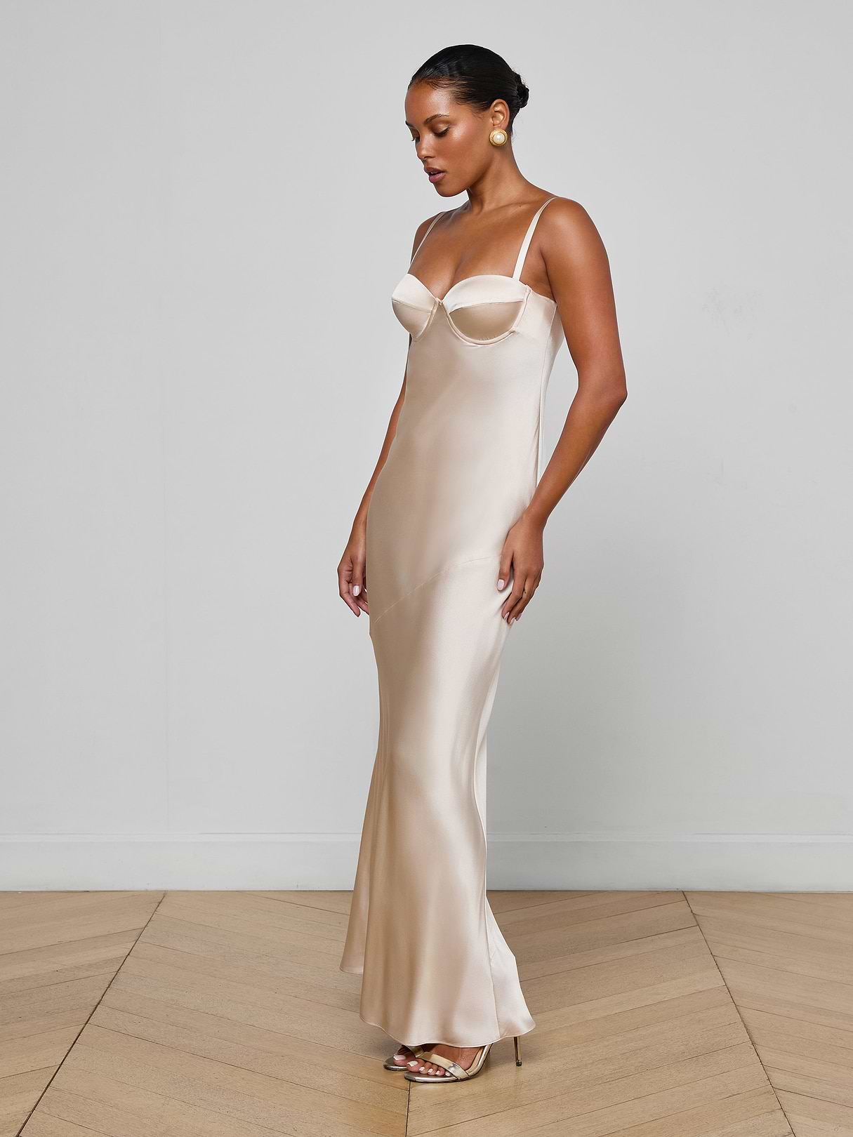 A woman in the Sayla Silk Gown—a long, light champagne dress with thin straps—stands against a plain white background in open-toe heels, looking down with a slight smile.