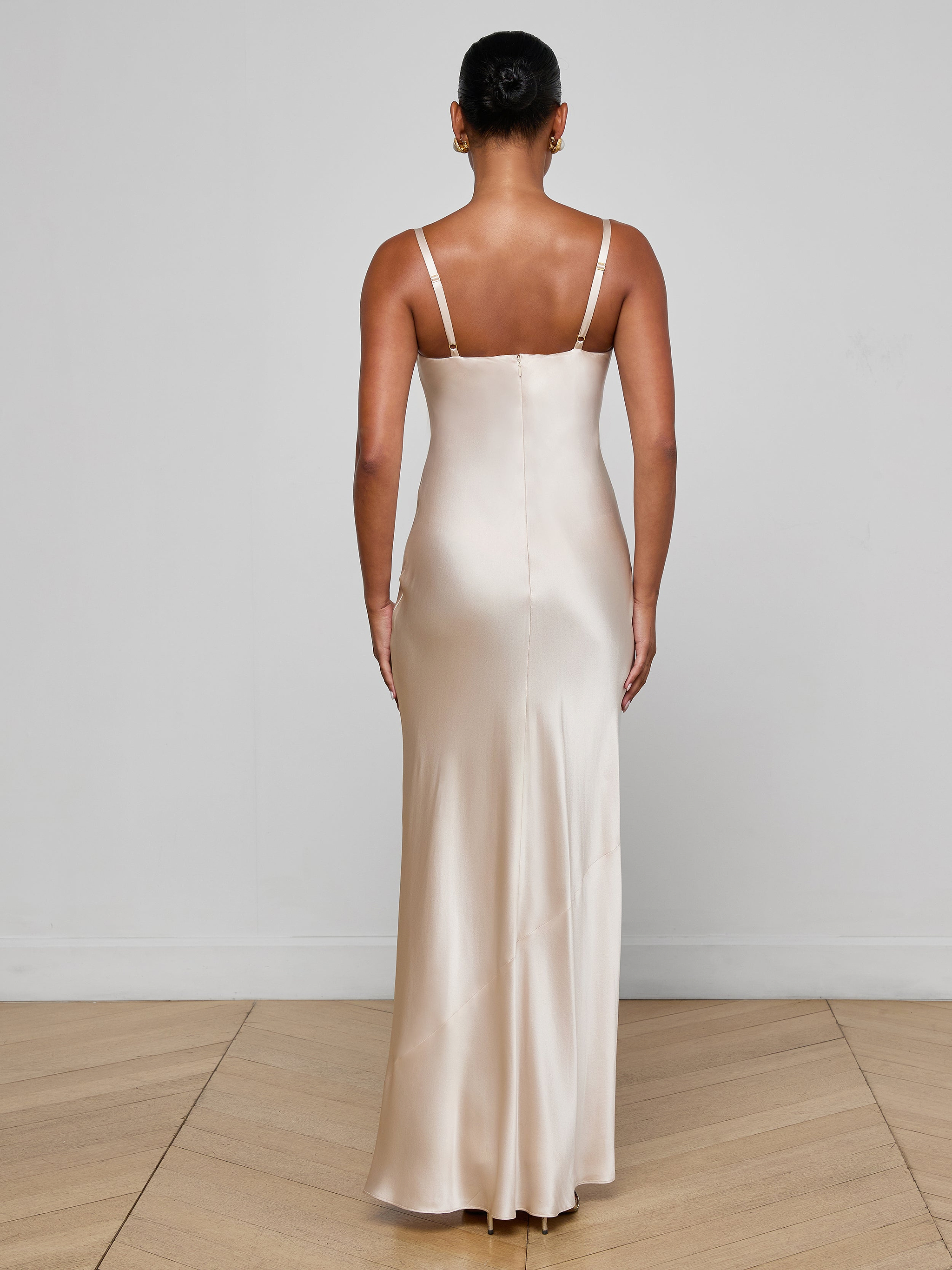 A woman stands with her back to the camera, wearing the Sayla Silk Gown—a long, sleeveless champagne dress with thin straps that exudes vintage glamour against a plain white background.