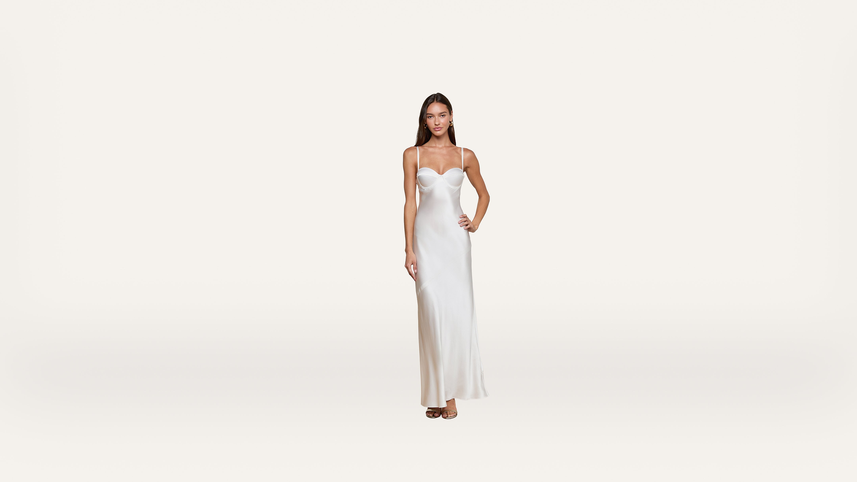 A woman stands wearing a long, white, sleeveless dress with thin straps. She has one hand on her hip and a neutral expression, set against a plain white background.