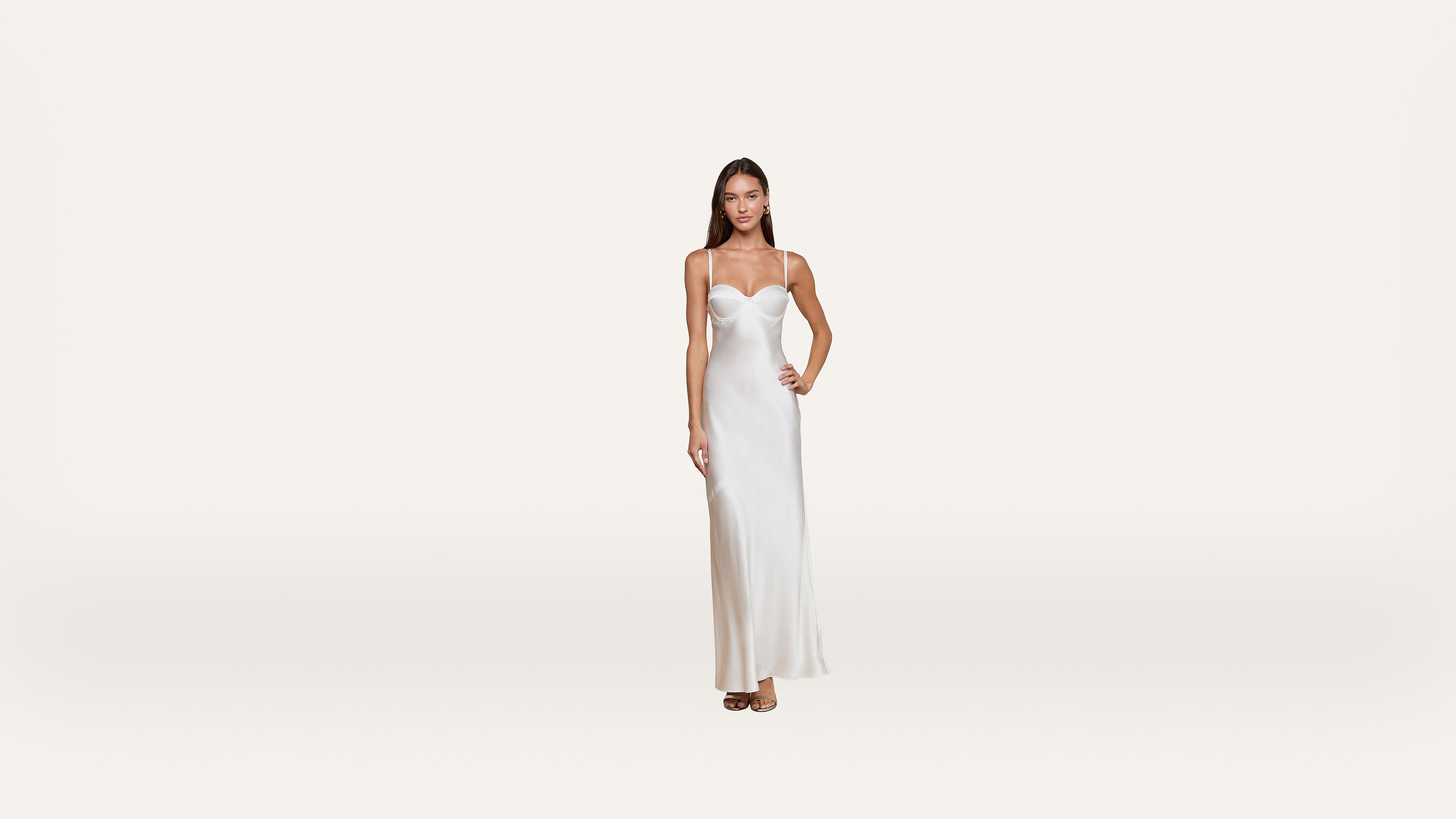 A woman stands wearing a long, white, sleeveless dress with thin straps. She has one hand on her hip and a neutral expression, set against a plain white background.