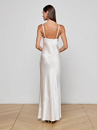 A woman stands facing away, wearing the Sayla Silk Gown—a long, white silk dress with thin straps that exudes vintage glamour. She has straight brown hair and large earrings, set against a plain white background.