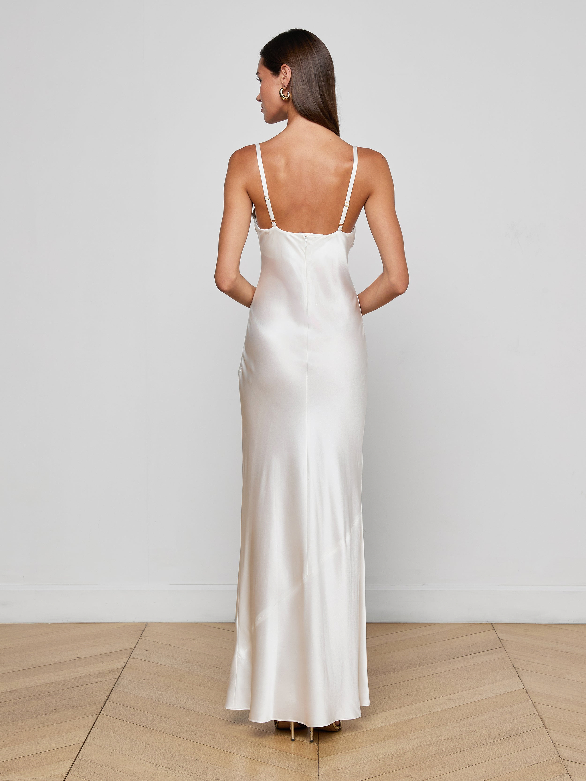 A woman stands facing away, wearing the Sayla Silk Gown—a long, white silk dress with thin straps that exudes vintage glamour. She has straight brown hair and large earrings, set against a plain white background.