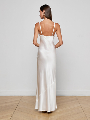 A woman with straight brown hair faces away, wearing the Sayla Silk Gown—a long, white silk dress with thin straps and an open back—perfect for events, styled on a light wood floor against a plain white wall.