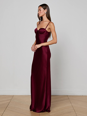 A woman stands sideways in the Sayla Silk Gown by L'AGENCE, featuring thin straps, a form-fitting bodice, and a flowing maroon skirt, radiating vintage glamour against a plain light gray wall and wooden floor.