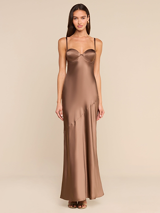 Against a beige backdrop, a woman exudes vintage glamour in the Sayla Silk Gown—a long, bronze satin slip dress featuring thin straps, a fitted bodice, and a flowing skirt. Her look is completed with long, straight hair and minimal accessories.