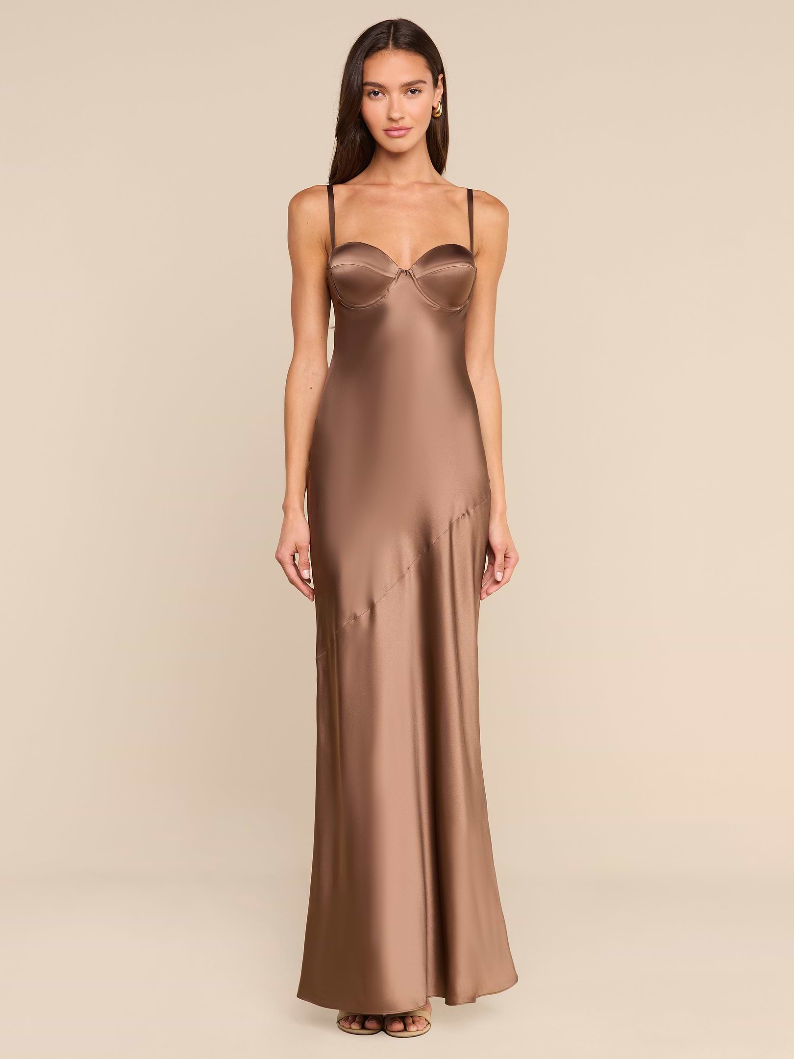 A woman stands facing forward in a plain, white studio setting, wearing the Sayla Silk Gown—a long bronze dress with thin straps.