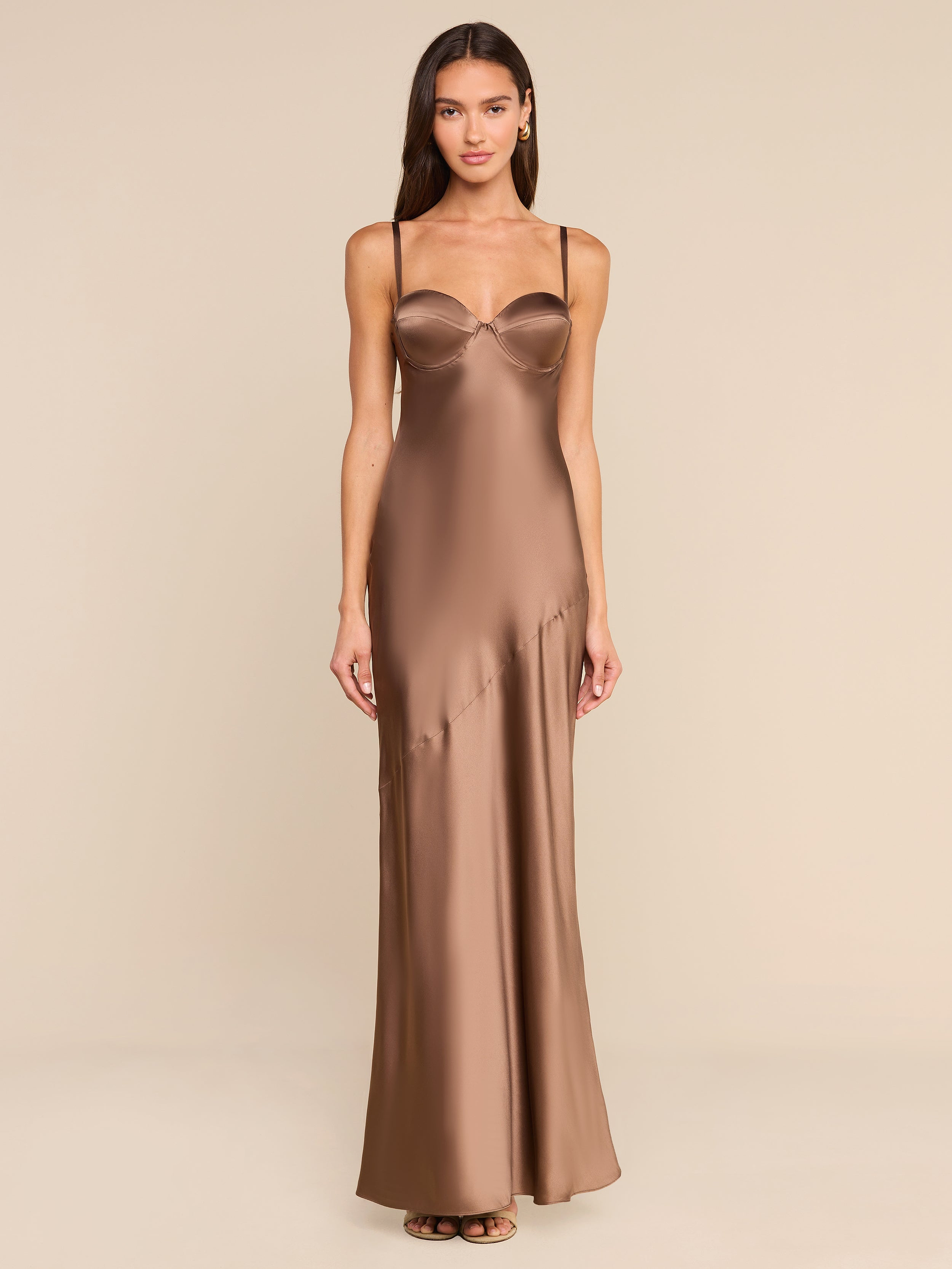 A woman stands facing forward in a plain, white studio setting, wearing the Sayla Silk Gown—a long bronze dress with thin straps.