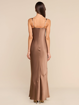 A woman stands facing away, exuding vintage glamour in the Sayla Silk Gown—a long, silky brown dress with thin straps—paired with beige heels against a plain light background.