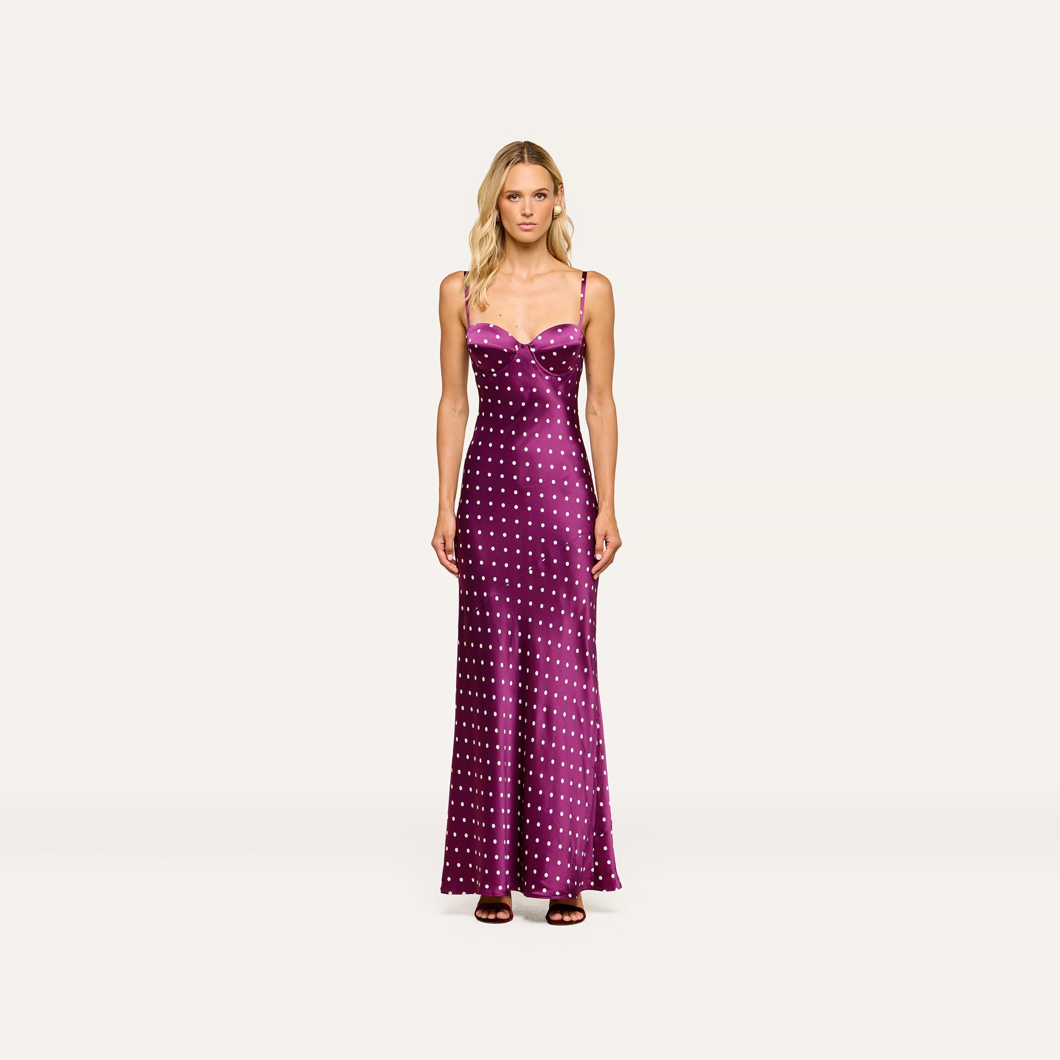 A woman stands facing forward against a plain light background, wearing the Sayla Silk Gown—a long, purple satin dress with white polka dots and spaghetti straps that exudes vintage glamour—paired with brown sandals.