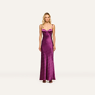 A woman stands facing forward against a plain light background, wearing the Sayla Silk Gown—a long, purple satin dress with white polka dots and spaghetti straps that exudes vintage glamour—paired with brown sandals.