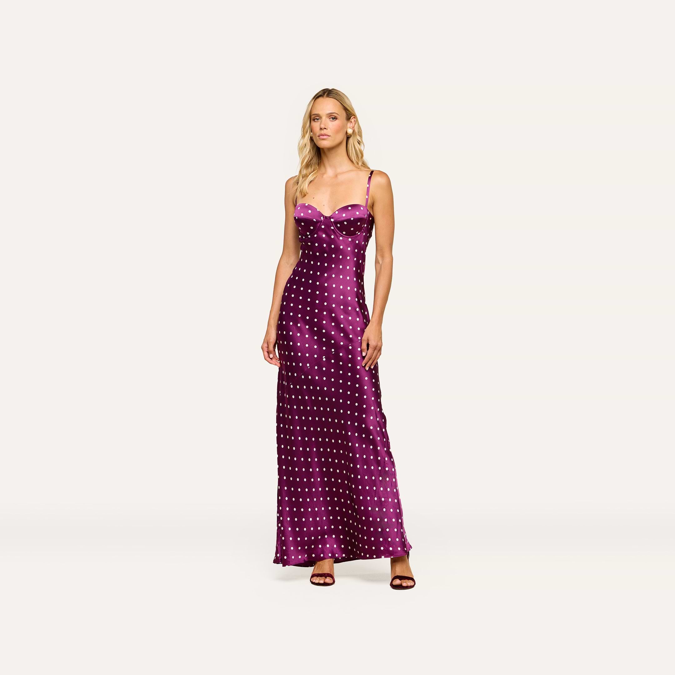 A woman stands wearing the Sayla Silk Gown, a long purple dress with white polka dots and delicate spaghetti straps, paired with dark heels against a plain light background, exuding vintage glamour.