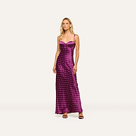 A woman stands wearing the Sayla Silk Gown, a long purple dress with white polka dots and delicate spaghetti straps, paired with dark heels against a plain light background, exuding vintage glamour.