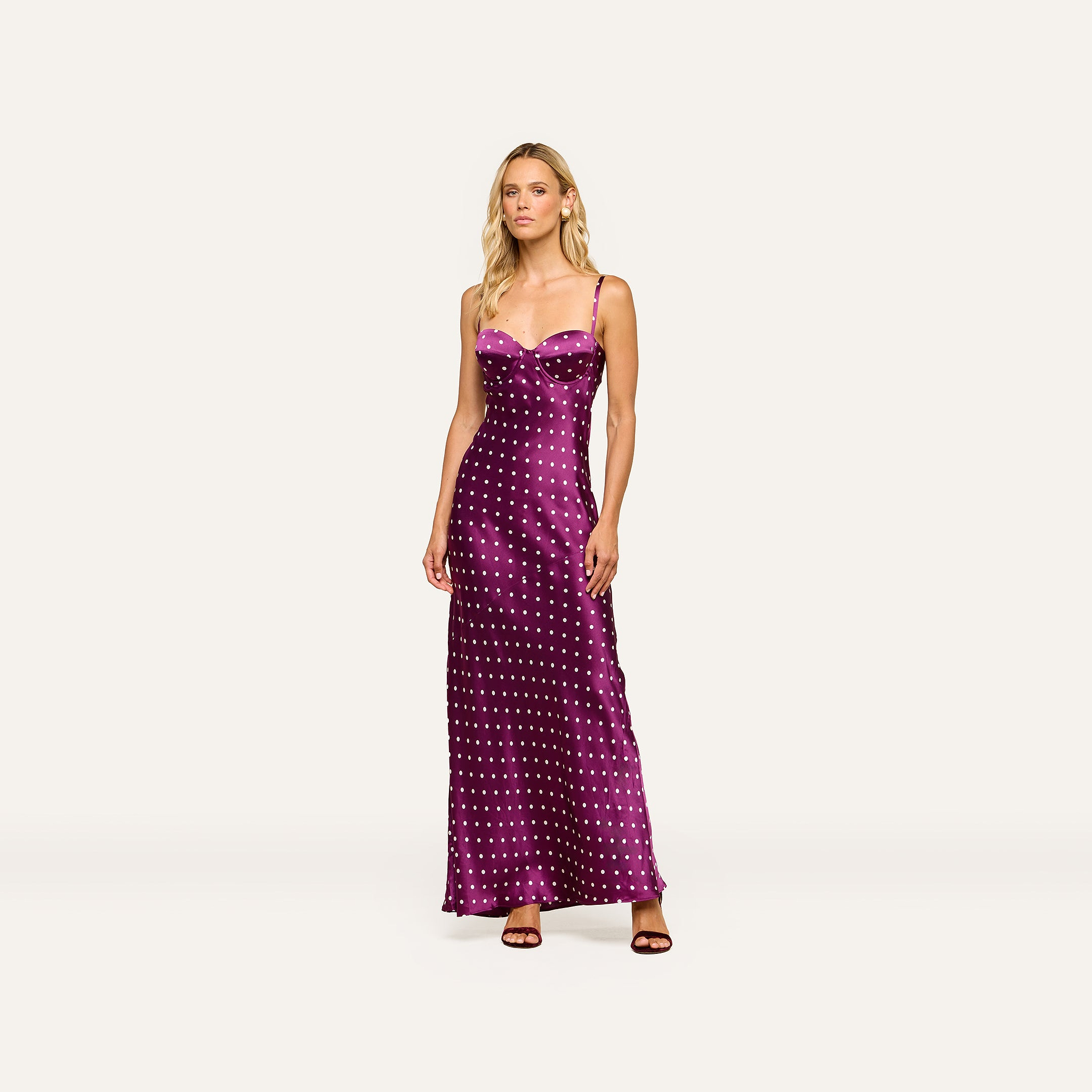 A woman stands wearing the Sayla Silk Gown, a long purple dress with white polka dots and delicate spaghetti straps, paired with dark heels against a plain light background, exuding vintage glamour.