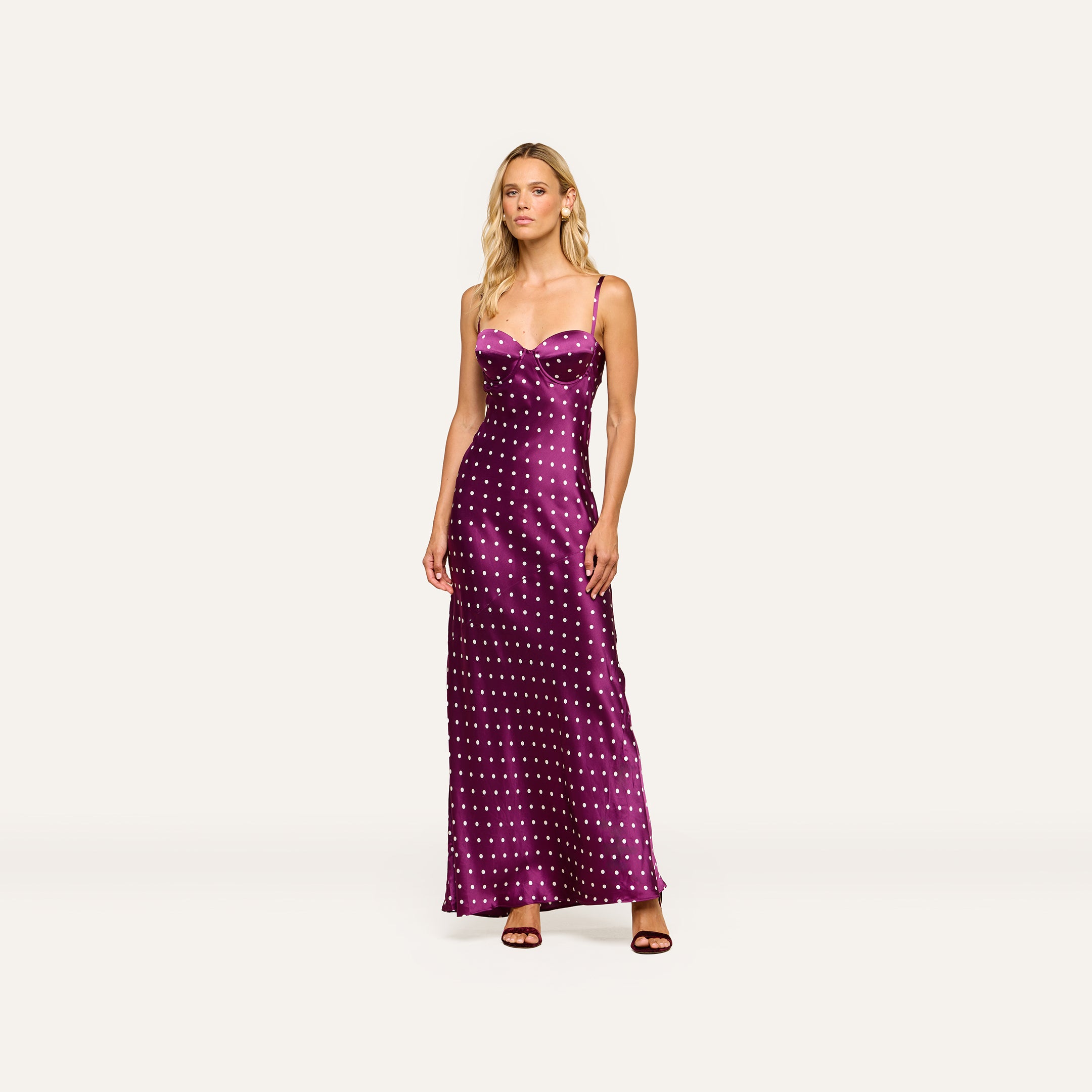 A woman stands wearing the Sayla Silk Gown, a long purple dress with white polka dots and delicate spaghetti straps, paired with dark heels against a plain light background, exuding vintage glamour.