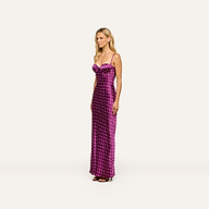 A woman stands against a plain, light background, exuding vintage glamour in the Sayla Silk Gown—an elegant, long purple polka-dot dress with spaghetti straps—paired with black heels.