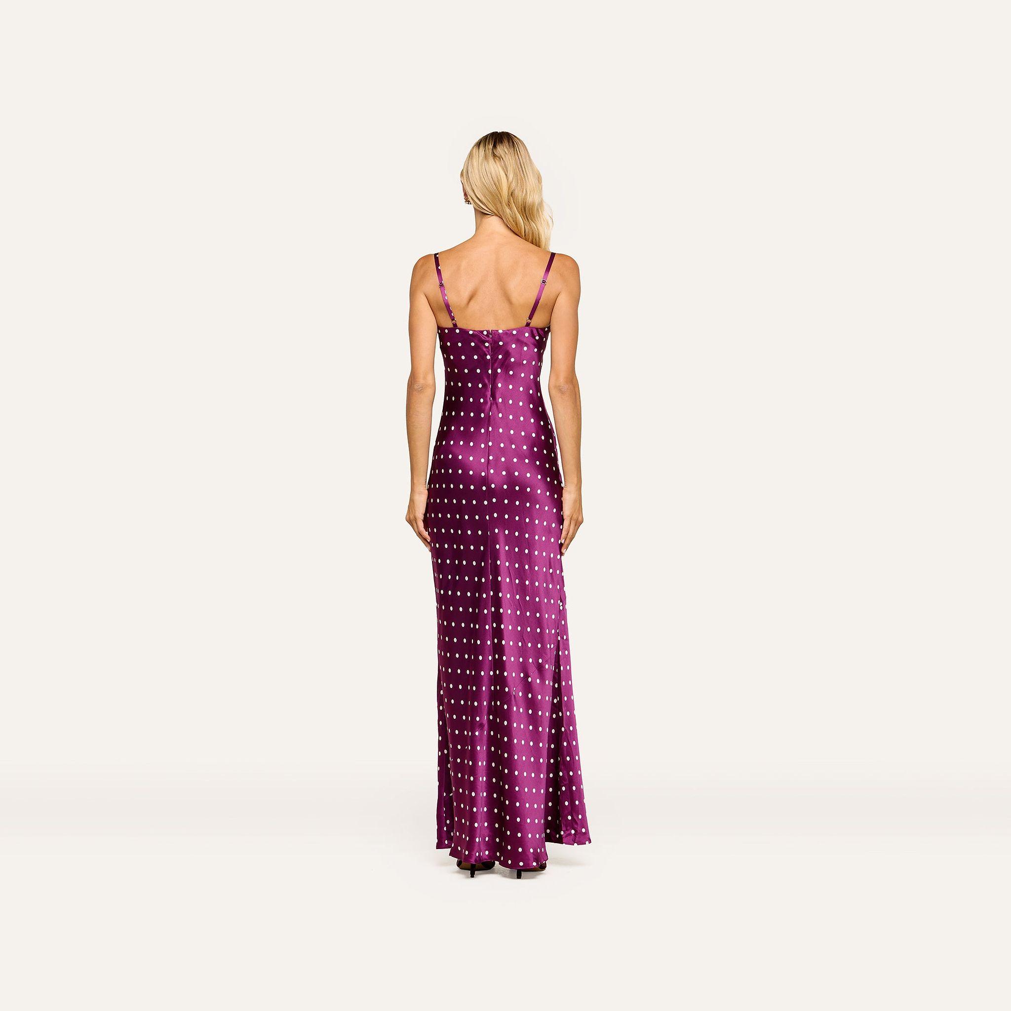 A person with long blonde hair is seen from the back, standing against a plain background, wearing the Sayla Silk Gown—a sleeveless, floor-length purple dress adorned with white polka dots, perfect for special events.