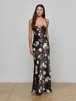 A woman with long brown hair poses against a plain light gray wall, wearing the L'AGENCE Sayla Silk Gown—a long black dress with thin straps and a yellow floral print—on a wooden floor.