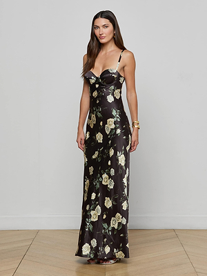 A woman with long brown hair wears the Sayla Silk Gown by L'AGENCE—a black, floor-length satin dress with light yellow rose prints, thin straps, and a sweetheart neckline. She stands indoors on a light wood floor against a gray wall.