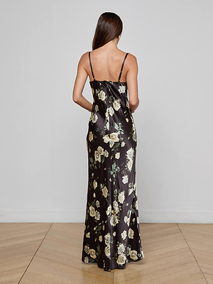 A woman with long brown hair, seen from behind, wears the L'AGENCE Sayla Silk Gown—a floor-length black satin dress with white and yellow floral prints and thin straps—standing against a plain light gray wall and wooden floor.