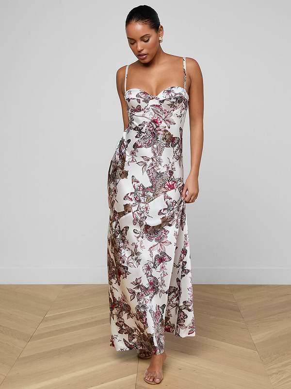 A woman stands barefoot on a wooden floor against a light gray background, wearing the Sayla Silk Gown—a long white dress with thin straps and a pink and black butterfly jungle print.