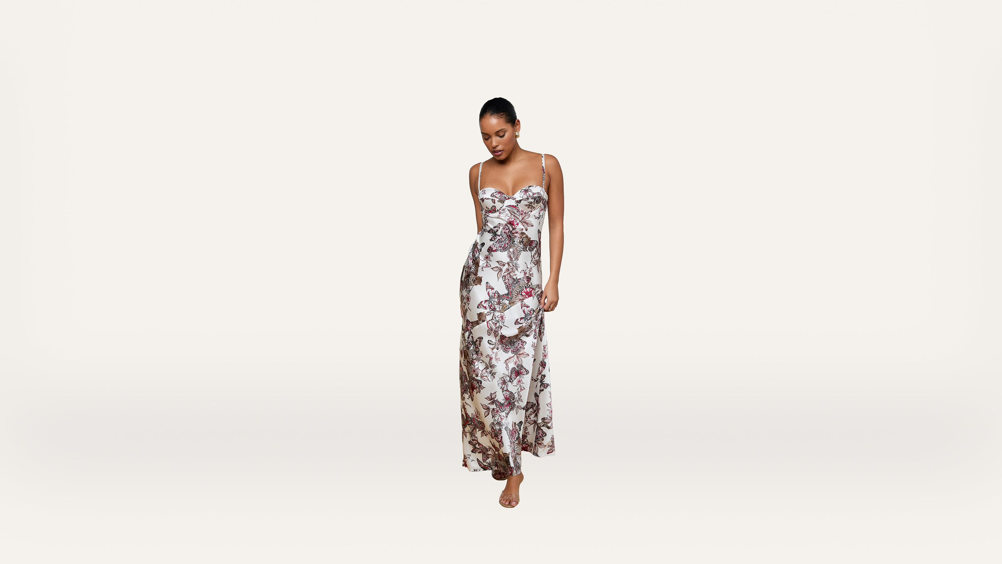 Woman in a long, sleeveless floral dress stands barefoot against a plain white background, looking down with her hands in her pockets.