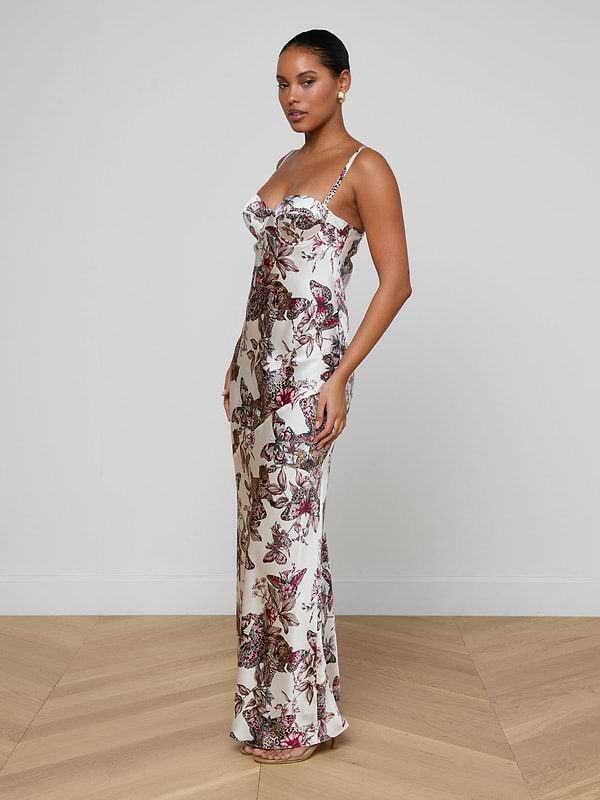 A woman stands indoors on a wooden floor, wearing the Sayla Silk Gown—a long, event-ready white dress with a floral butterfly jungle print and thin straps. She faces sideways to the camera, her hair pulled back, arms relaxed at her sides.