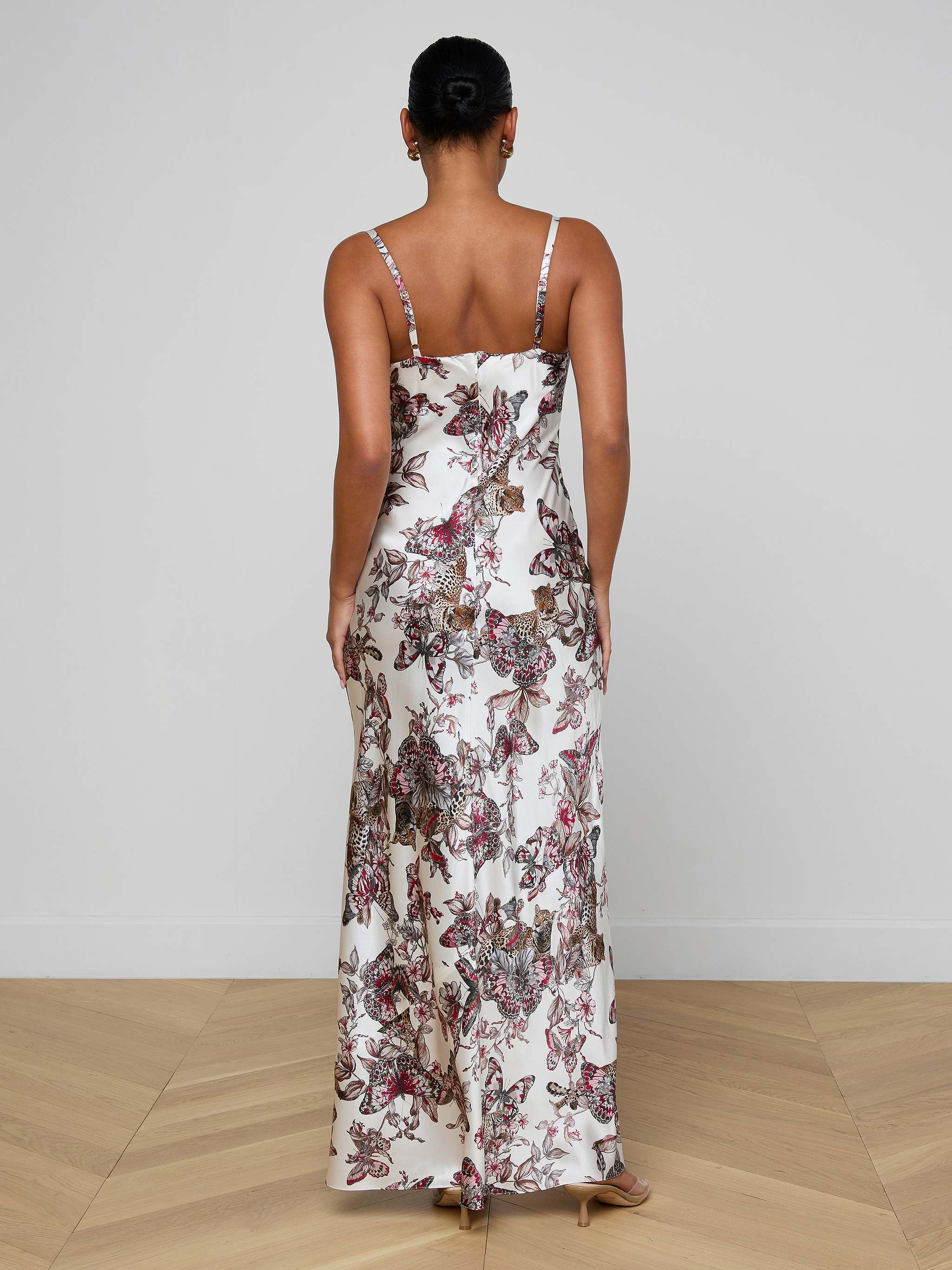 A woman stands on a light wood floor against a white wall wearing the Sayla Silk Gown, event-ready with a flowing skirt, thin straps, and a striking butterfly jungle print.