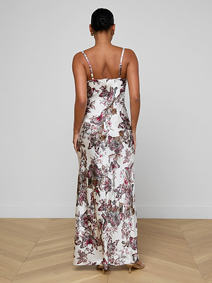 A woman stands on a light wood floor against a white wall wearing the Sayla Silk Gown, event-ready with a flowing skirt, thin straps, and a striking butterfly jungle print.
